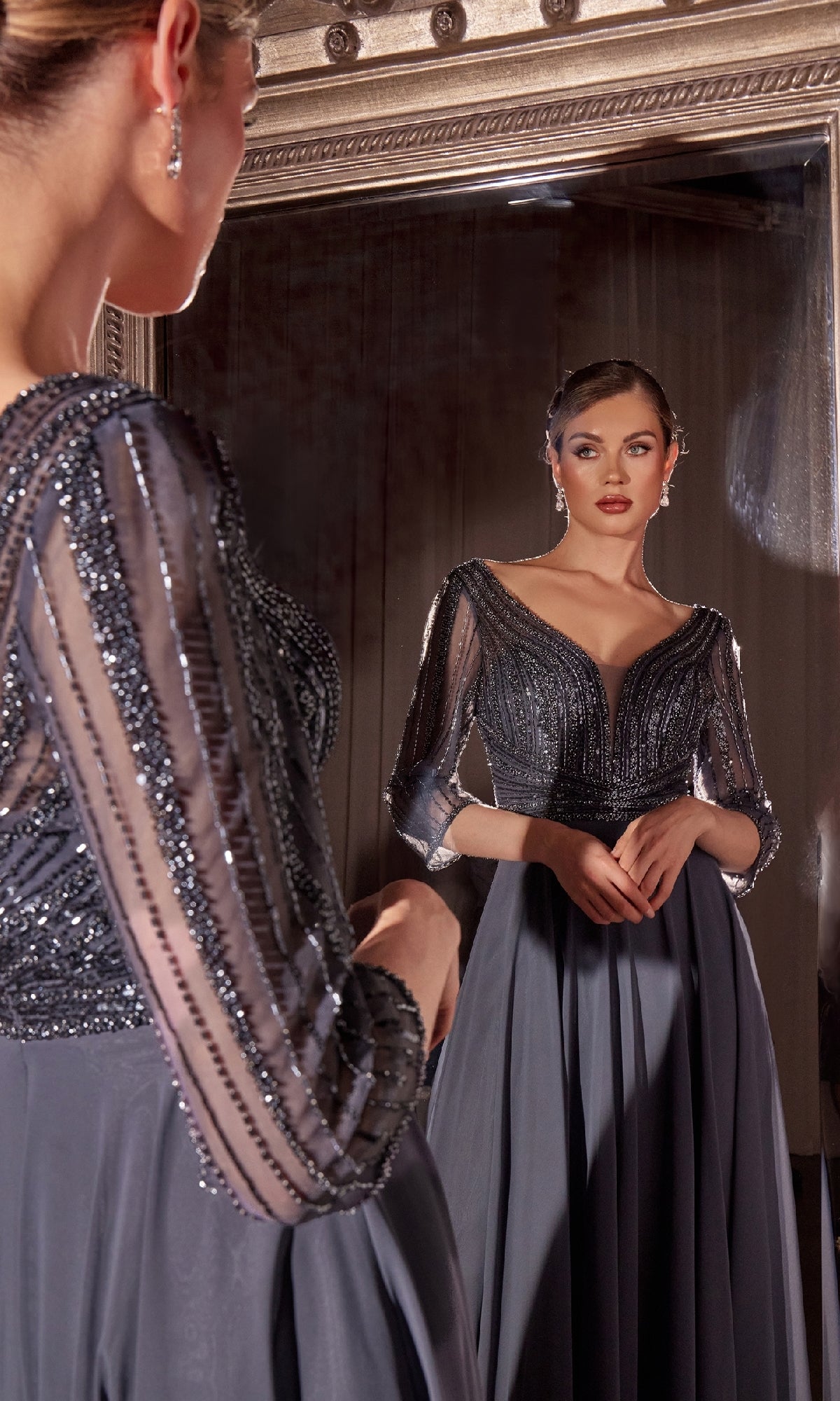 A woman in the Ladivine CC446 Modest Long A-Line Formal Dress with sheer beaded sleeves stands before a large mirror, gracefully admiring her reflection.
